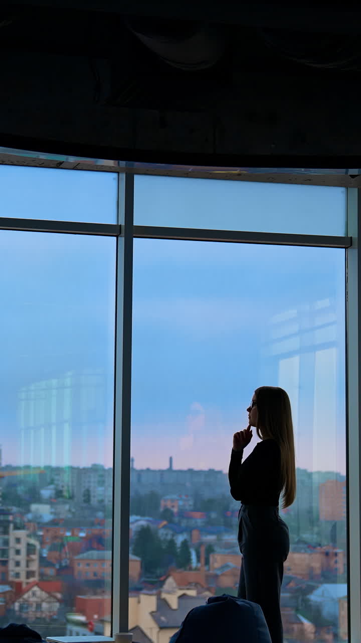Young business woman thinking near the window. Silhouette of a pretty lady in office in the evening. Panoramic window office view with a city landscape. Vertical video