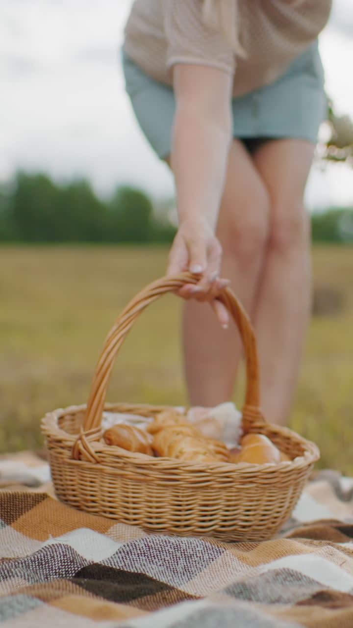 una manta de picnic a cuadros en un campo de hierba mientras una persona coloca suavemente una canasta tejida llena de bocadillos horneados, la suave luz dorada del sol destaca el campo rural con balas de heno en la distancia