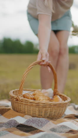 checkered picnic blanket on grassy field as person gently places woven basket filled with baked snacks, soft golden sunlight highlights rural countryside with hay bales in distance
