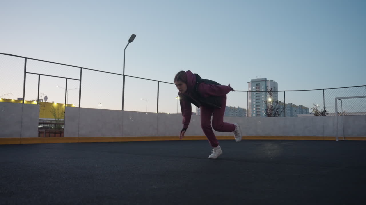 Female runner on white court line getting set to run at dusk on black asphalt sports court near white barrier topped with chain link fence in urban setting wearing maroon hoodie and white sneakers