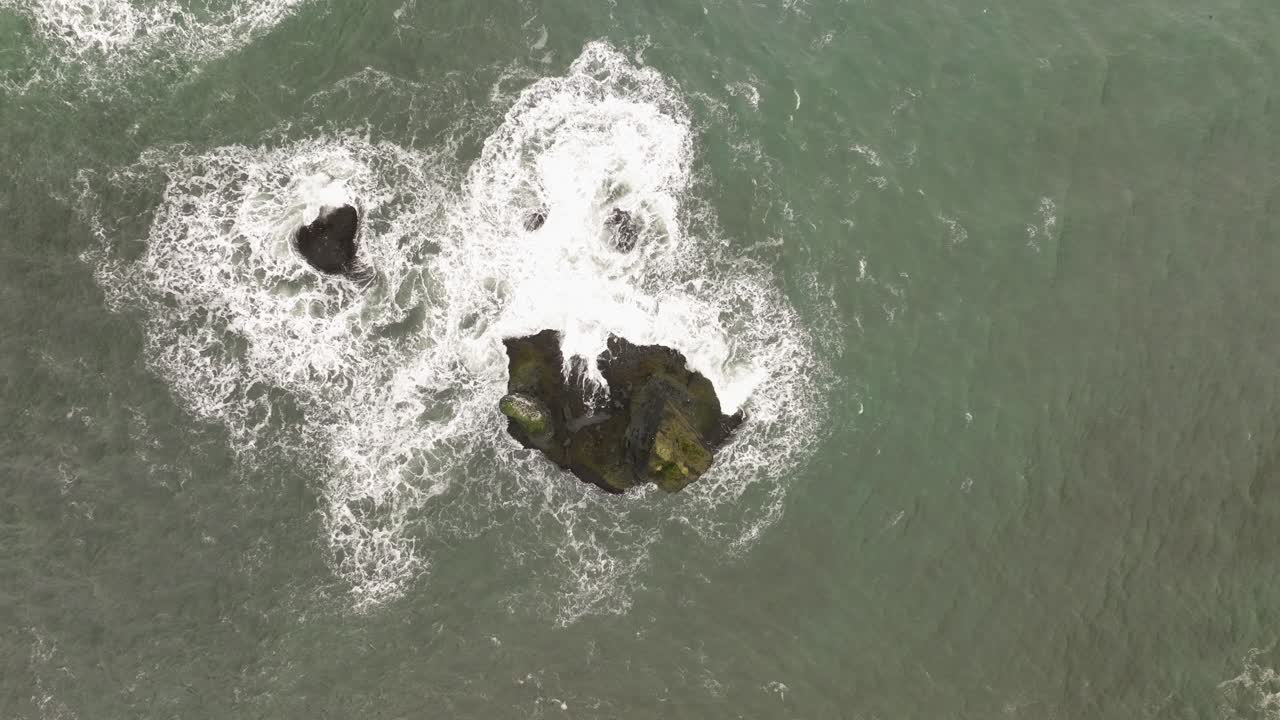 Aerial view of isolated rock formations in the ocean at Reynisfjara, Iceland, surrounded by crashing waves, highlighting the rugged volcanic coastline.