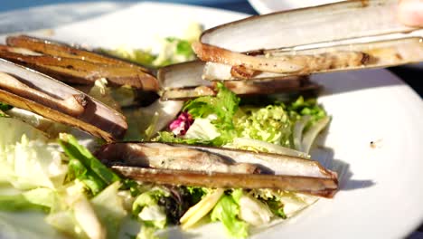 Tomando-A-Mano-Almejas-A-La-Parrilla-En-Un-Plato-Blanco-Con-Lechuga.