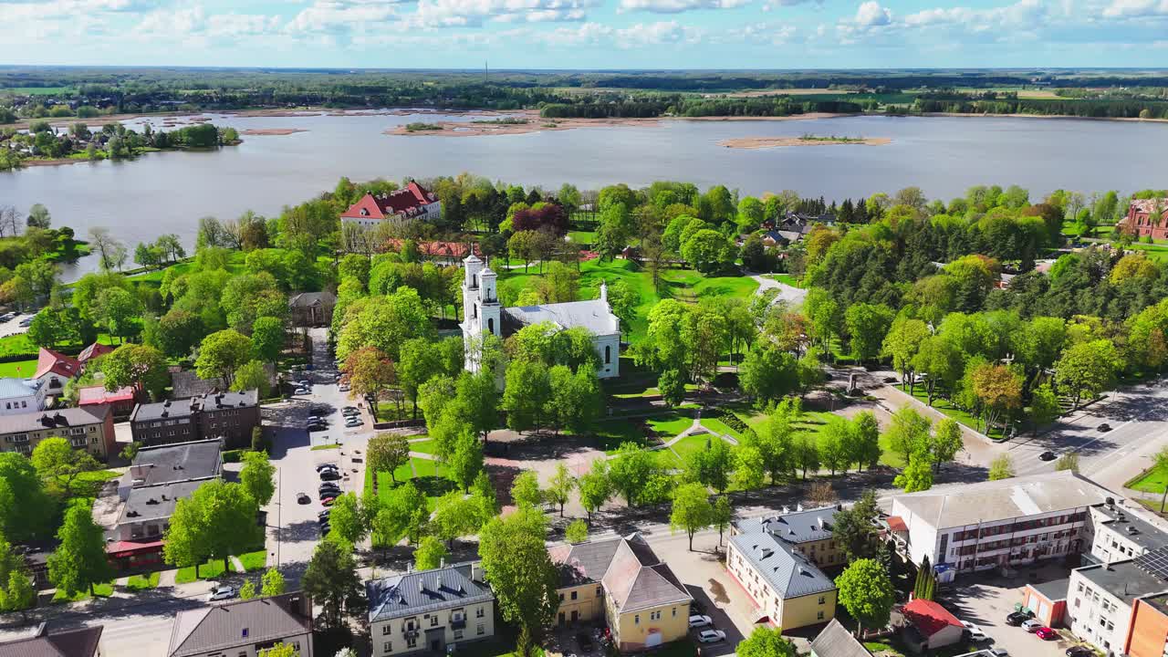 Aerial view of a scenic riverside town featuring a white twin-tower church, residential buildings, lush parks, and calm water with islands under a blue sky.
