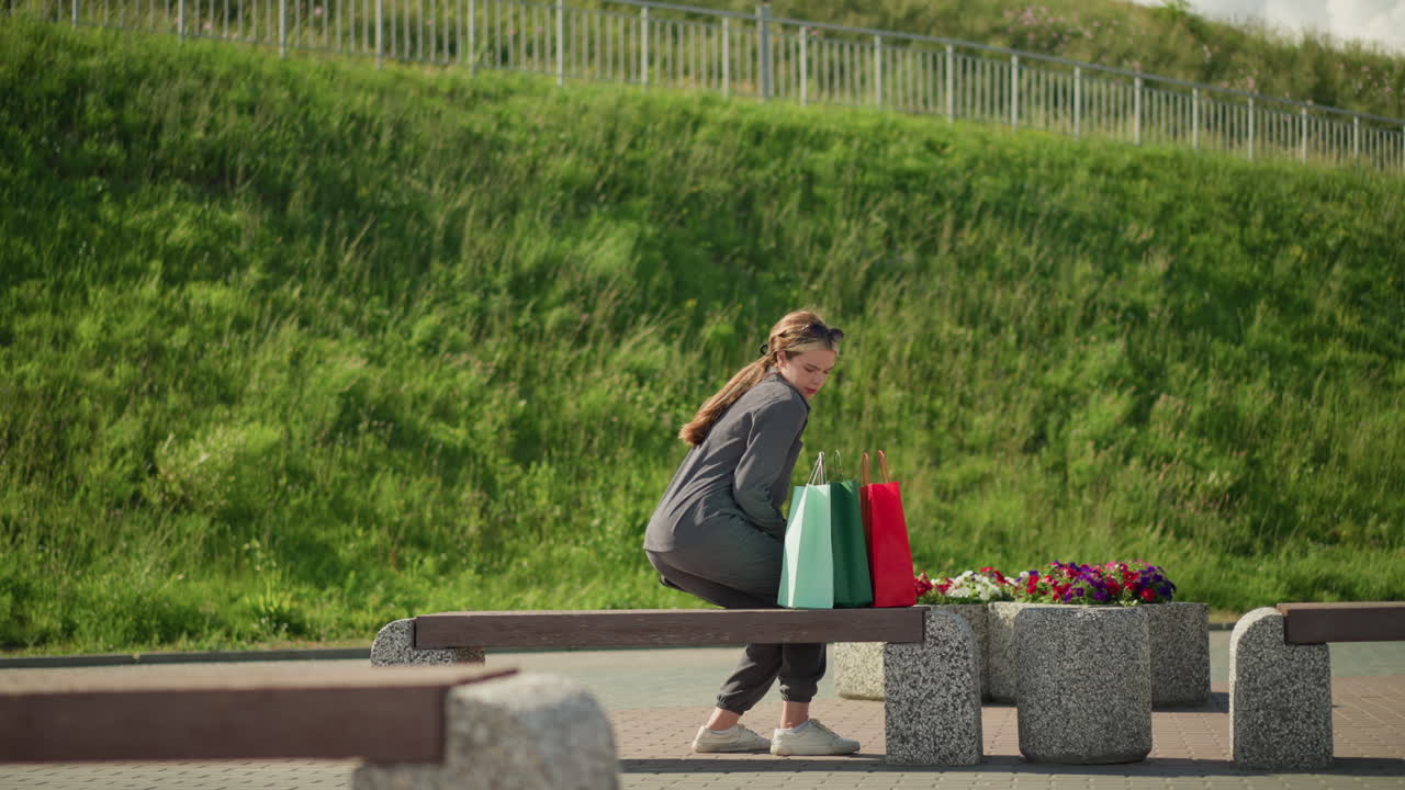 여자는 벤치에 걸어, 다채로운 쇼핑 가방을 떨어뜨리고, 좌석에 앉아, 왼손을 벤치 위에 놓고, 다리를 조정하고, 멀리, 초록색과 뒷면의 언덕에서 볼 수 있는 빛의 기둥을 쳐다보고 있습니다.