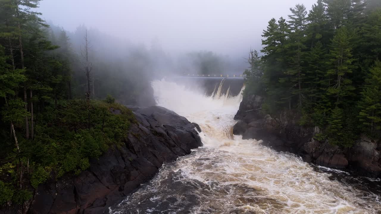 A powerful waterfall spilling from a dam, surrounded by rocky cliffs and dense green forests under a misty atmosphere.