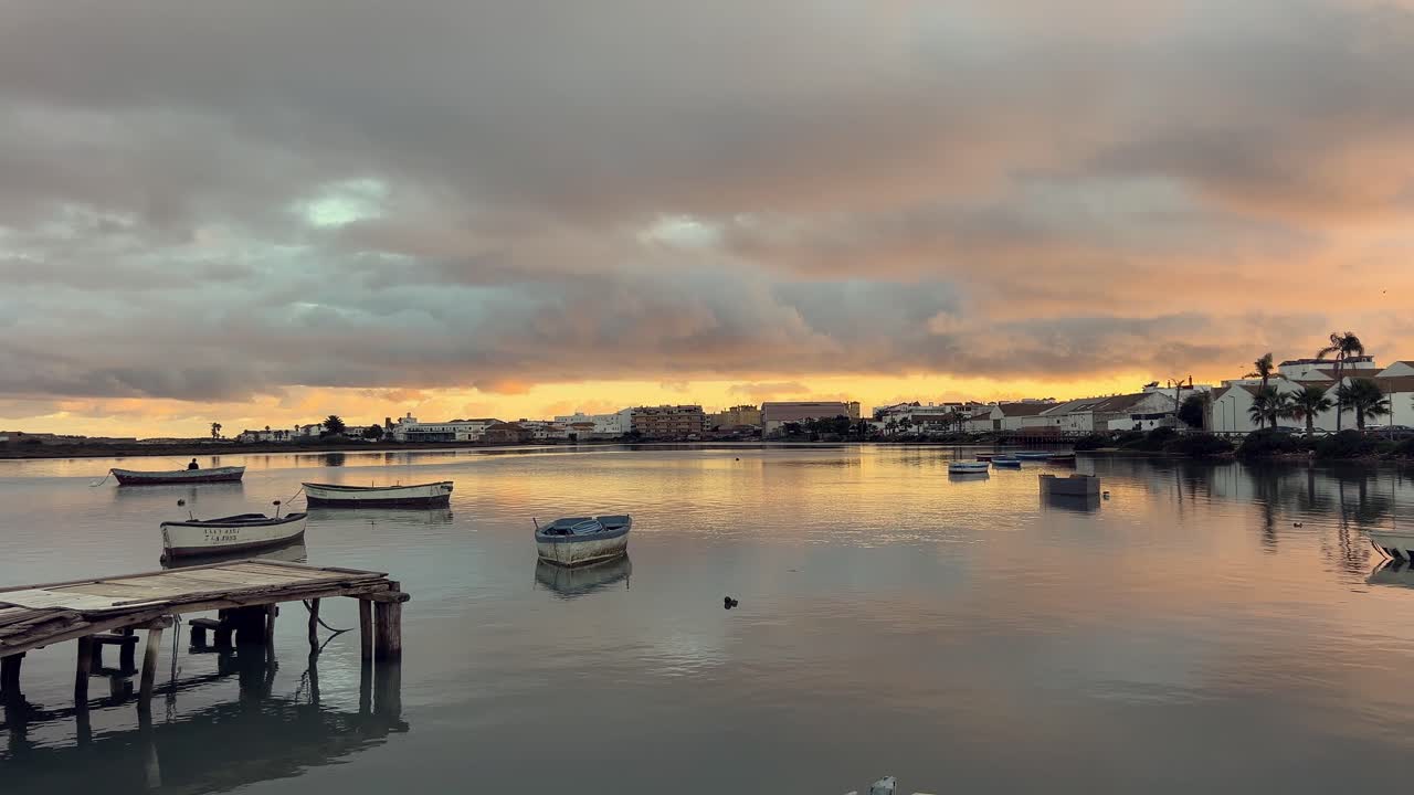 스페인 안달루시아에 있는 바르바테 항구에 조용한 해가 뜨고, 평온한 바다에서 부드럽게 흔들리는 어선들이 다채로운 하늘을 반사하여 평온한 해안 풍경을 만니다.