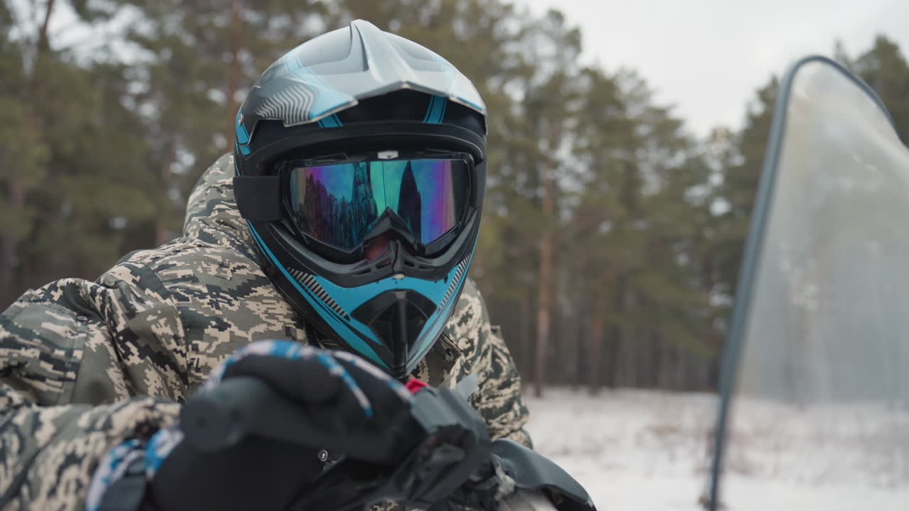 ATV-instruktør tester gasspedalen på en snødekt løype, kamuflasjekledd fører med blå hjelm og reflekterende briller øver på kontroll og balanse, overfører kraft gjennom styret, skog i bakgrunnen, overskyet himmel i fokus.