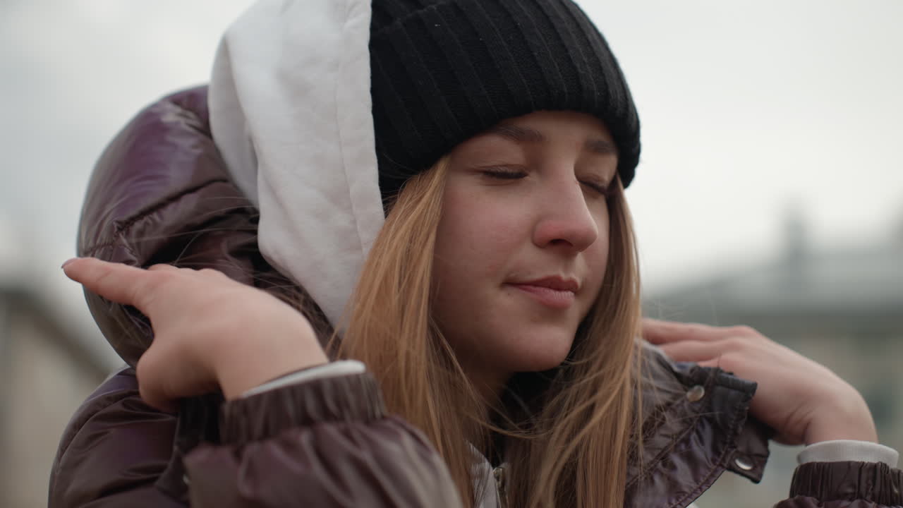 Женщина стоит возле домов в ветреный день, на ней шапка бини и пуховик, она смотрит на городской ветер, случайный снимок женщины в холодную погоду, поправляющей капюшон при сильном ветре и мрачном небе