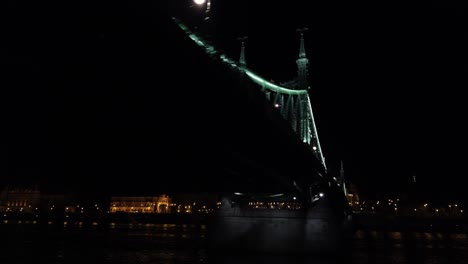 passing by liberty bridge in budapest, hungary, with the gellért hotel and hill with the citadella in the background on the other side of river danube at night