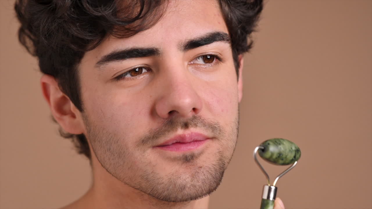 A young caucasian man with stubble beard is doing a facial massage using a roller. Slow motion