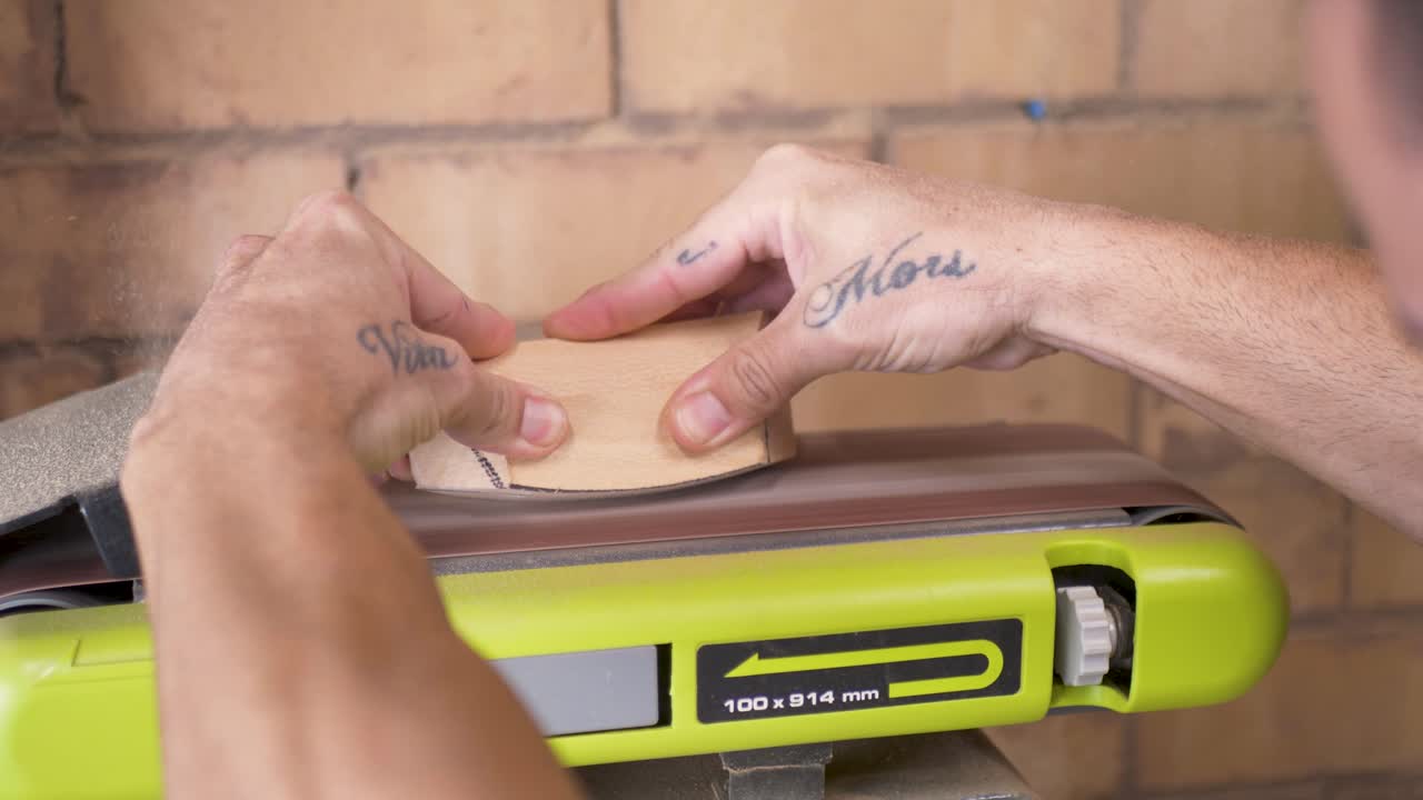 Slow motion close-up of hands holding a hardwood timber block for shaping climbing holds with electric belt sander tool in home garage workshop shed iAustralia carpentry project building industry