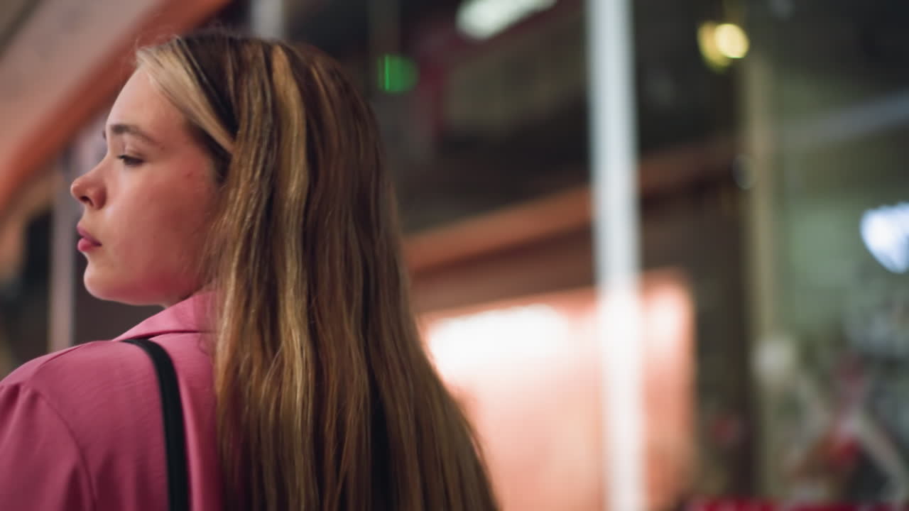 분홍색 드레스를 입은 여자가 검은 가방을 들고 밝게 밝은 쇼핑몰에서 흐릿한 배경으로 다른 사람들이 돌아다니는 것을 암시하며, 그녀의 표정은 그녀의 주변을 관찰하는 것처럼 평온합니다.