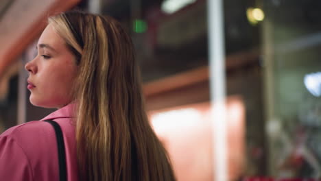 dama de vestido rosa sosteniendo una bolsa negra en un centro comercial brillantemente iluminado con un fondo borroso, sugiriendo otras personas caminando, su expresión es tranquila mientras parece observar su entorno
