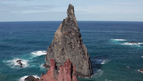 flying over mountain to reveal beautiful rock in vast blue ocean during sunny day, water in the horizon, waves rolling in creating foam