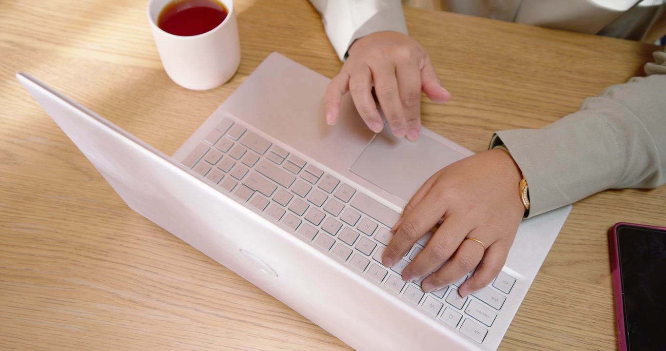 Mujer africana ajustando los dedos, tocando el trackpad y escribiendo en un portátil plateado en un escritorio con una taza y un teléfono.