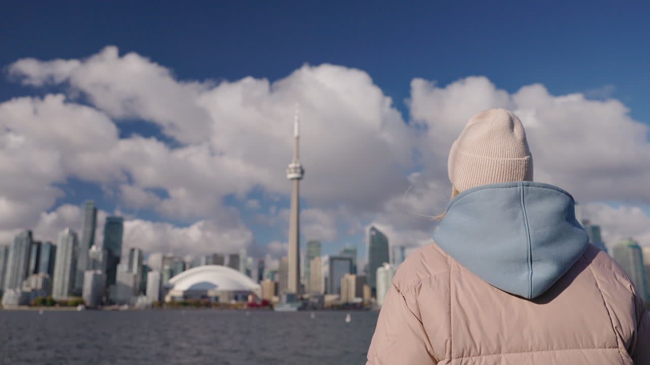 Giovane turista che osserva lo skyline di Toronto dal lungolago del Lago Ontario, un'adolescente di spalle con giacca calda e cappello si gode il panorama urbano e l'atmosfera di viaggio.