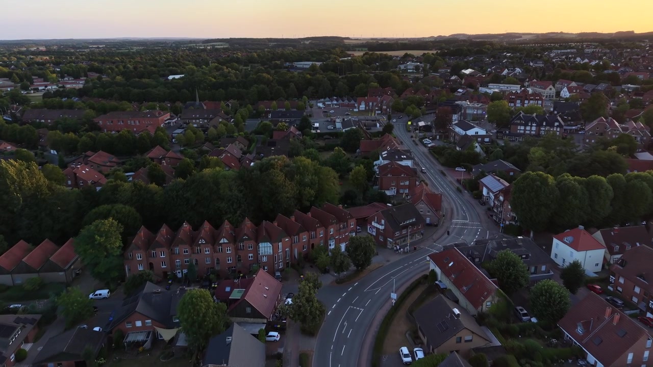 Case a schiera con tetti rossi costeggiano una strada curva attraverso una piccola città tedesca al tramonto, densi isolati residenziali si estendono verso campi lontani sotto una calda luce dorata serale. Ampia ripresa aerea da drone