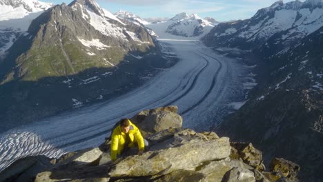 joven con chaqueta amarilla en formación rocosa, senderismo en rocas, con vistas al épico glaciar aletsch, el glaciar más grande de los alpes