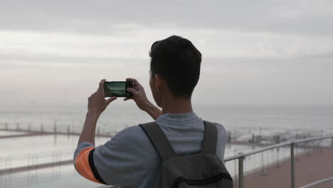 Retrato-De-Un-Joven-Asiático-Tomando-Una-Foto-De-Una-Playa-Nublada-Usando-El-Teléfono