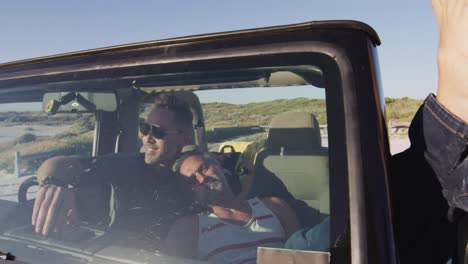 Happy-caucasian-gay-male-couple-in-car-relaxing-with-feet-out-of-window-on-sunny-day-at-the-beach