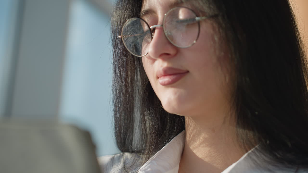 Extreme close up of young woman with glasses focused on tablet screen in sunlit indoor space, displaying deep concentration and serene expression as natural light highlights her face and hair