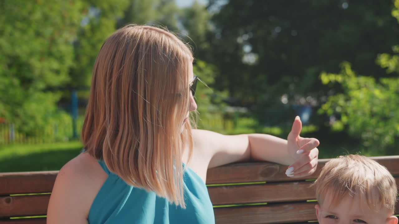 Mor og barn på bænk i solrig park, mor giver tommelfinger op til dreng, afslappet sommerkjole og solbriller, grønne træer og sollys, oprigtig samhørighed og blid samtale, weekendudflugtsstemning.