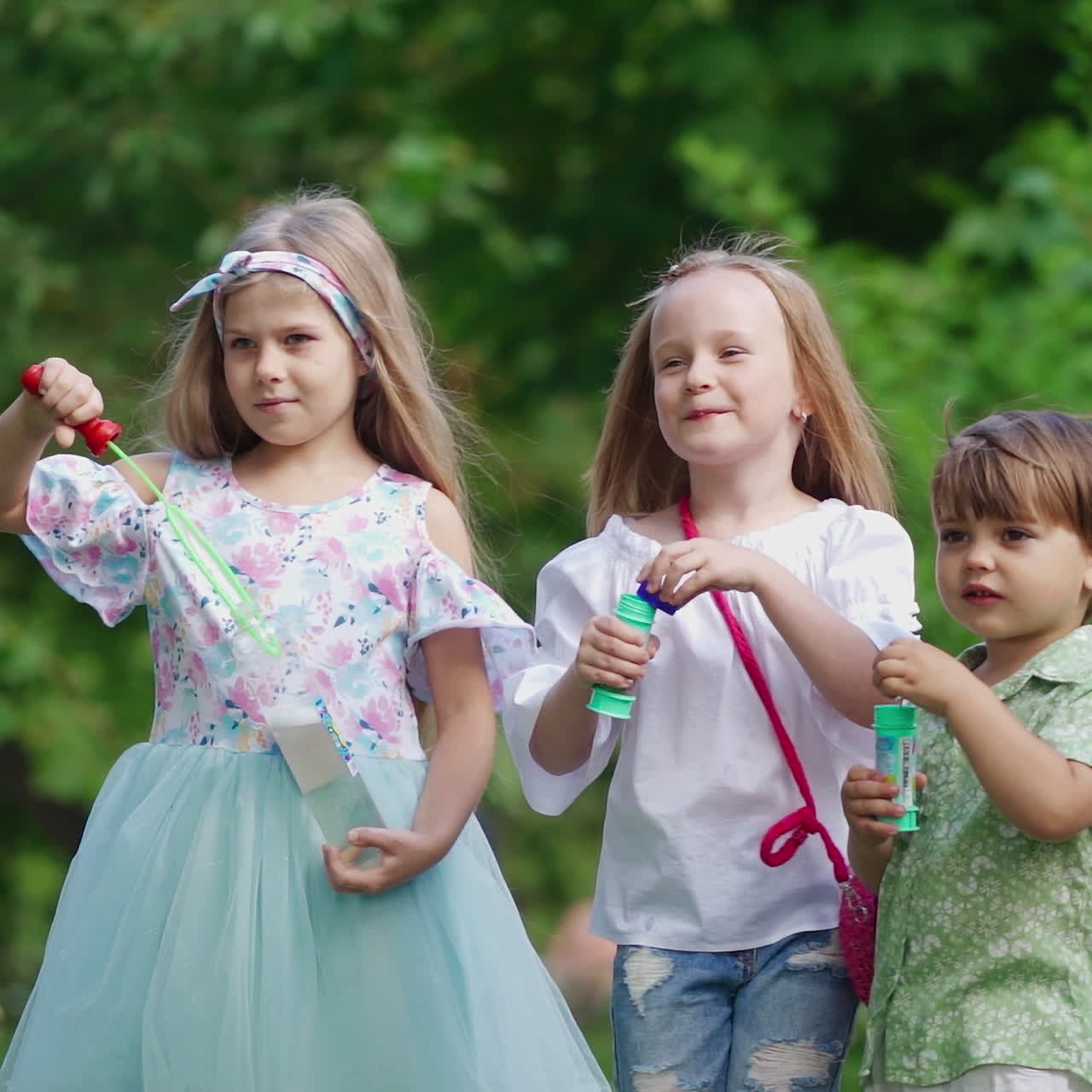 Happy kids have a rest in the park in a summer day. Little children blow air bubbles on the background of green bushes outdoors.
