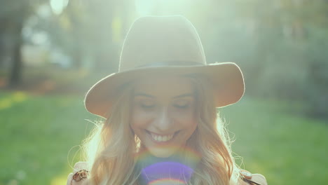 mujer rubia joven caucásica con un sombrero sonriendo y mirando a la cámara mientras sostiene hojas amarillas de otoño en el parque