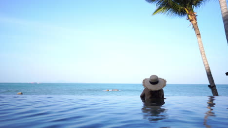A-woman-relaxing-in-an-infinity-swimming-pool,-during-the-day,-looking-out-at-the-ocean-view