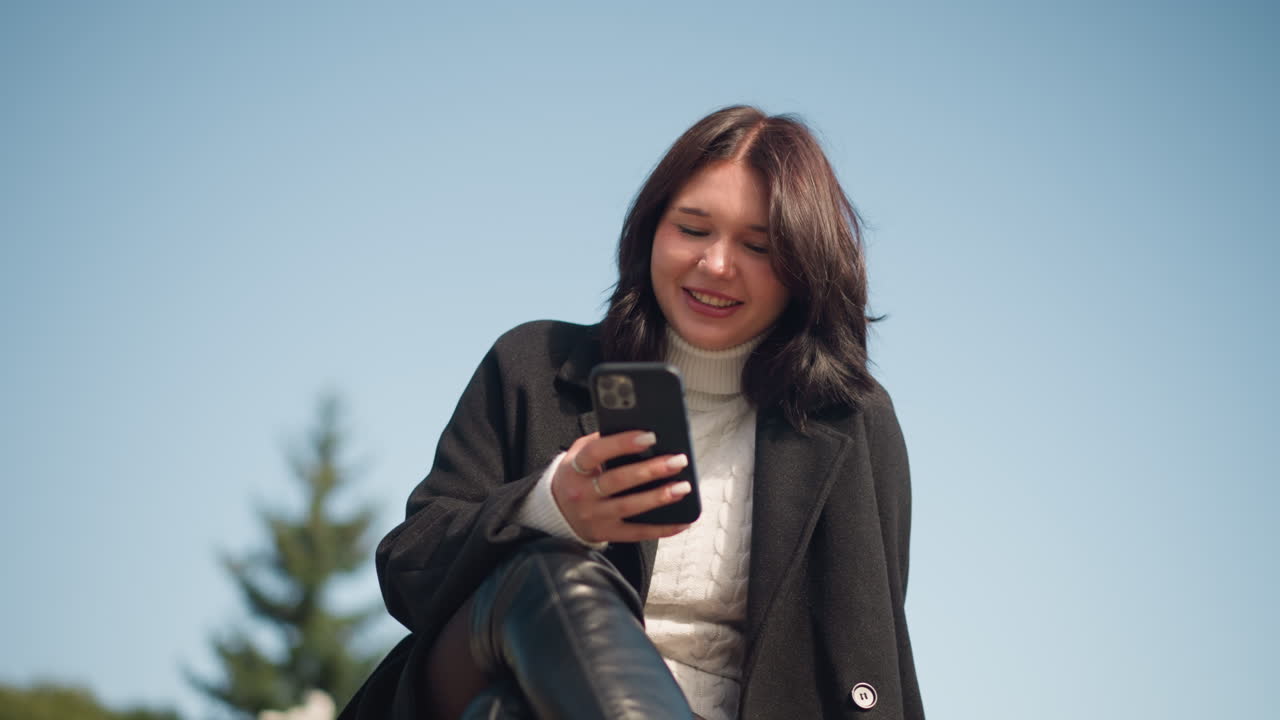 Professional lady smiling while focusing on smartphone in park setting, wearing black coat and white sweater, with legs crossed, enjoying serene outdoor atmosphere under blue sky and lush greenery