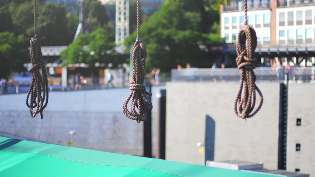 Tied marine ropes hanging from a boat with people walking around a harbour in the blurred background on a sunny day.