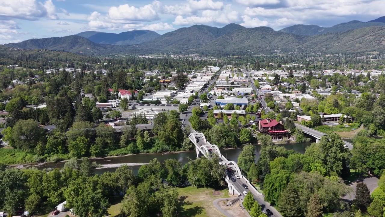 Skyline del centro di Grants Pass con il Ponte Caveman sul fiume Rogue, con alberi verdi e rigogliosi e le montagne dell'Oregon meridionale sullo sfondo. Ampia ripresa aerea con drone