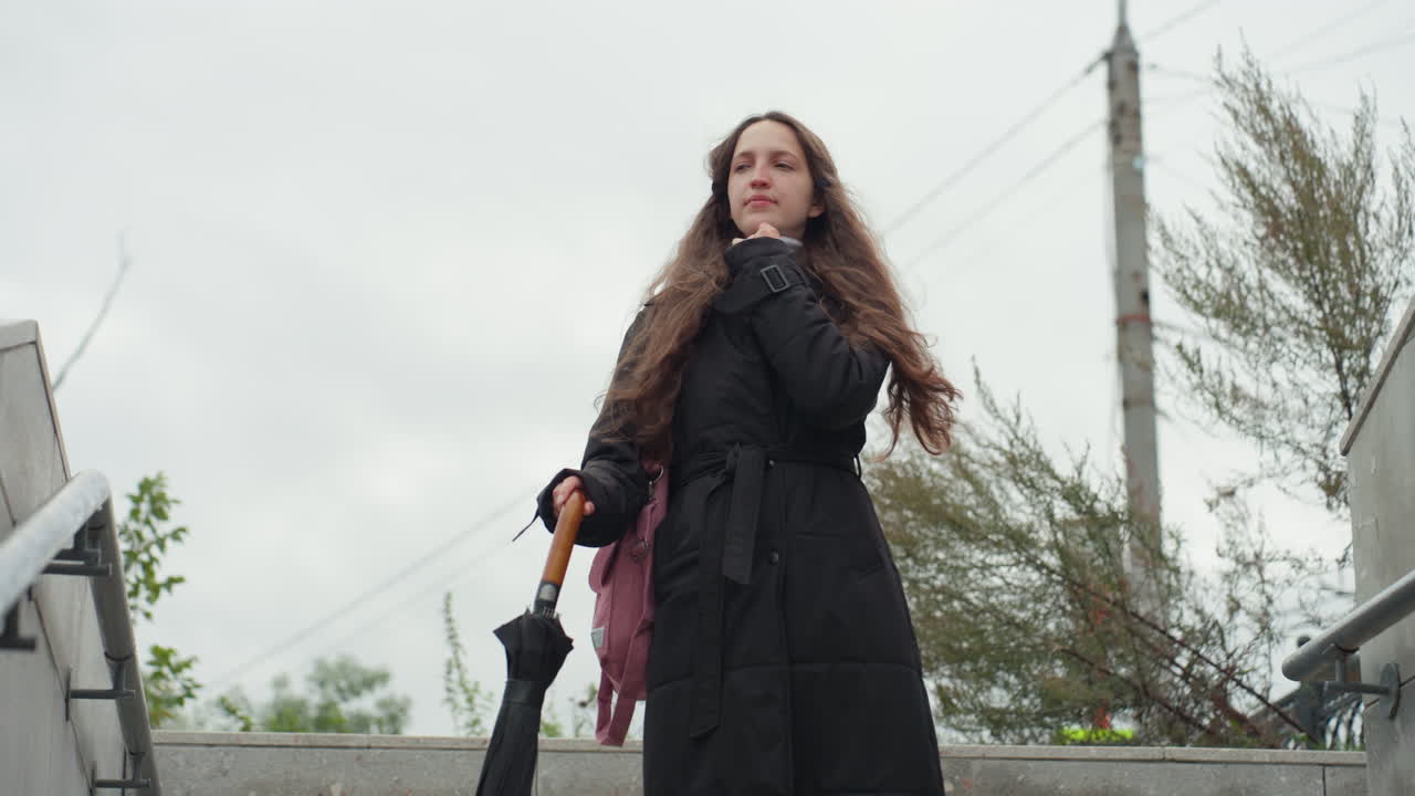 Donna che scende le scale di cemento con un cappotto nero, tiene in mano un ombrello e una borsa, lunghi capelli ricci mossi dal vento, cielo nuvoloso, sequenza di immagini di pendolare urbana che la mostrano come pendolare, studentessa, viaggiatrice