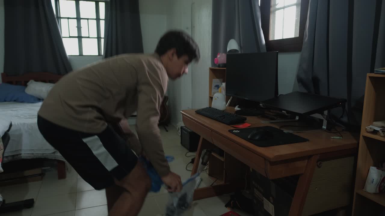 Young Caucasian man organizing and cleaning under messy desk in bedroom. Wide shot.