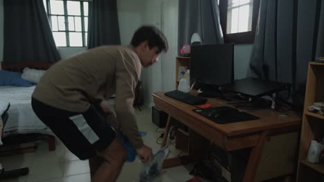 young caucasian man organizing and cleaning under messy desk in bedroom. wide shot.