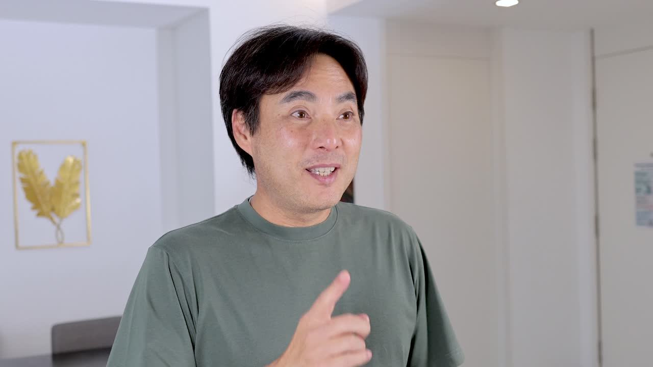 Asian man in casual shirt smiles, gesturing quiet with finger on lips indoors