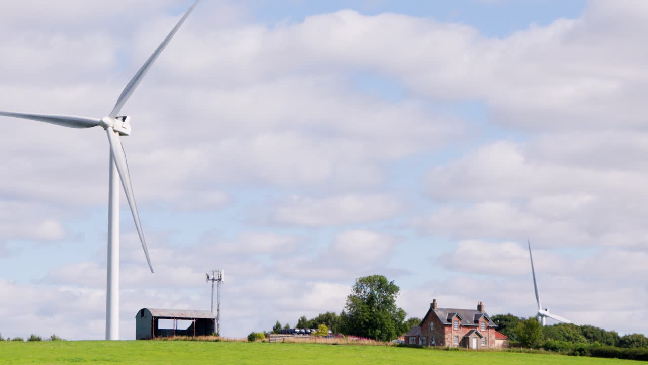 A large wind turbine slowly rotates beside a farmhouse and green fields under partly cloudy daylight in rural Edinburgh, Scotland. Static wide shot, natural lighting