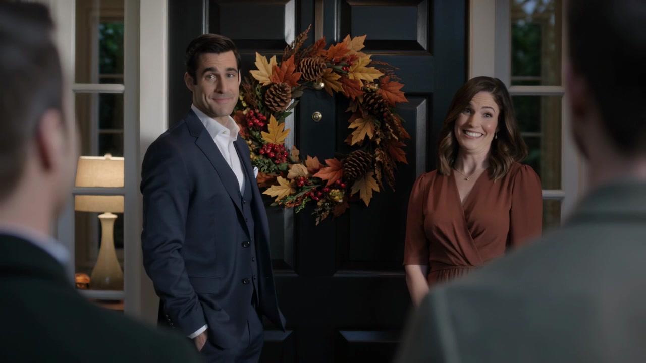 I padroni di casa, con abito e vestito color ruggine, sistemano una ghirlanda autunnale e accolgono gli ospiti in arrivo alla porta nera.