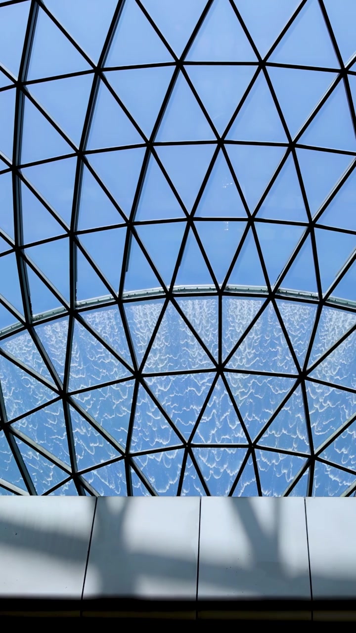 La telecamera si inclina verso l'alto sotto una cupola di vetro geometrica, rivelando una cascata interna circolare con la luce naturale del giorno che illumina la struttura architettonica.