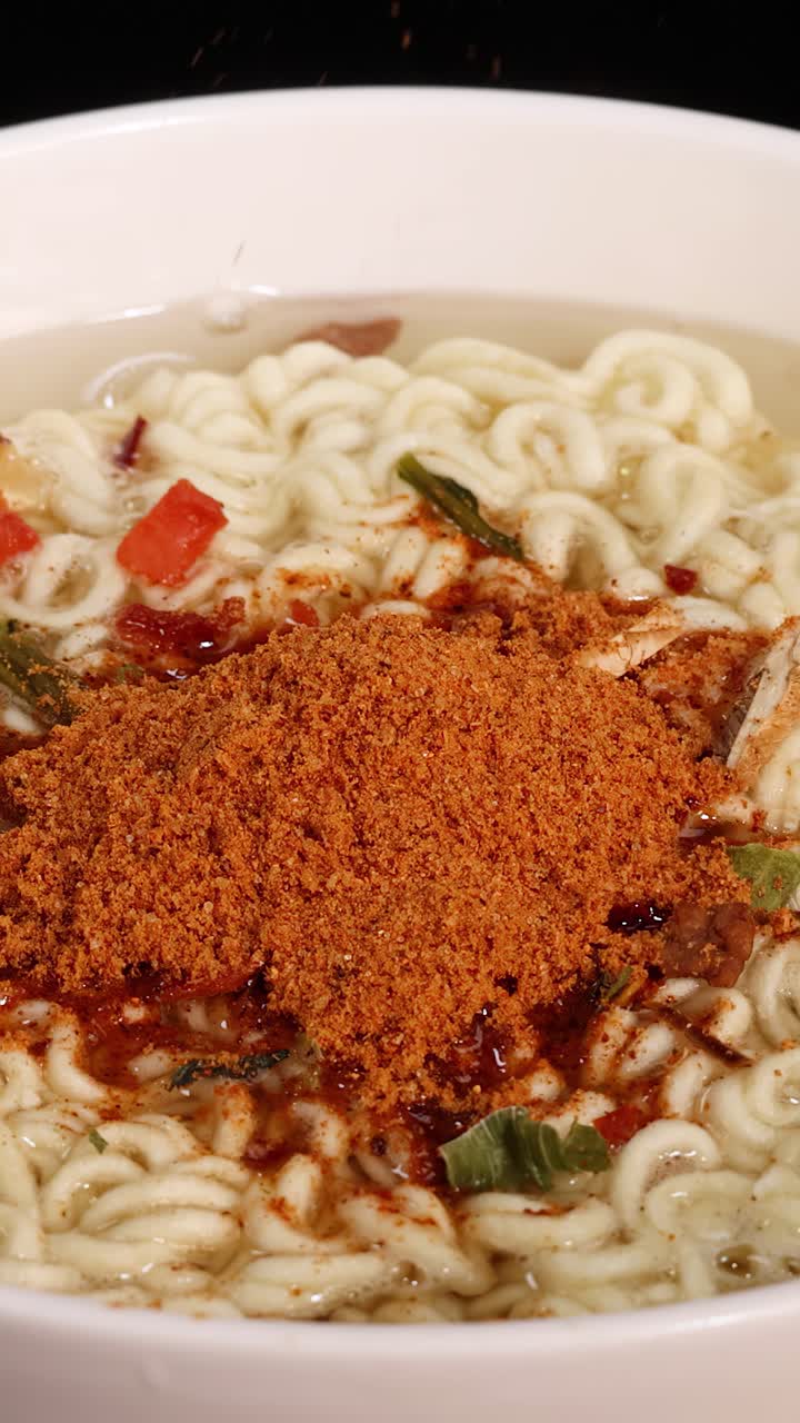 明るいスタジオ照明の下、白い器に入った乾燥野菜入りの熱いインスタントラーメンに、粉末スパイスミックスが振りかけられるクローズアップ。
