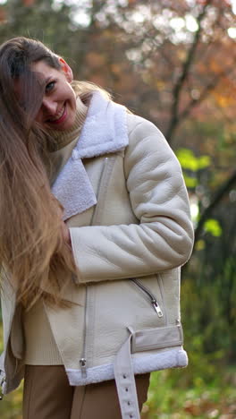 smiling caucasian lady in white jacket waving her long hair. woman having fun relaxing in the walk at autumn park. blurred backdrop. vertical video