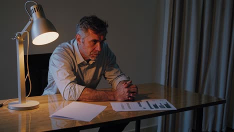 professional businessman analyzing financial documents and graphs, sitting at desk in dimly lit office, experiencing work related stress during late evening hours