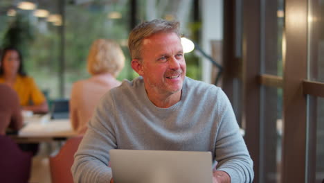 Mature-Businessman-Working-On-Laptop-At-Desk-In-Office-Pausing-To-Look-Out-Of-Window