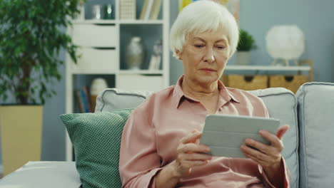 Gray-Haired-Senior-Woman-Sitting-On-The-Couch-With-Tablet-Device-And-Her-Cute-Teenage-Granddaughter-Coming-From-Behind-And-Hugging-Her