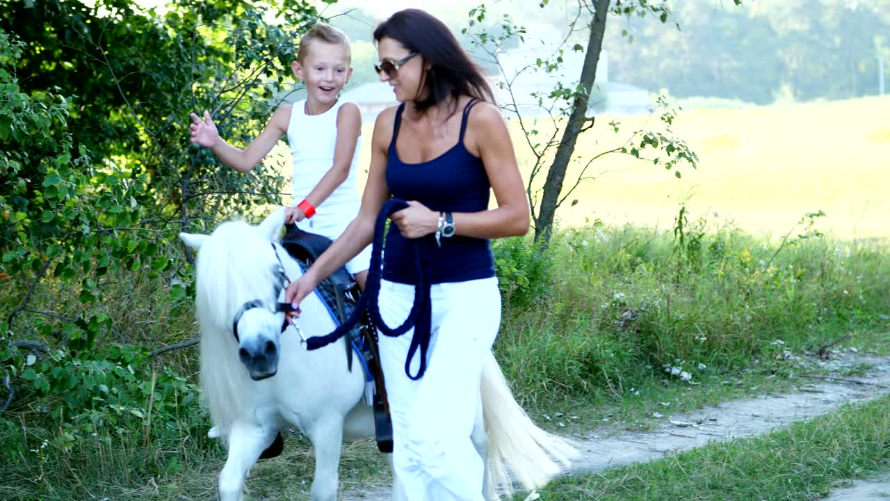 A woman and a boy are walking around the field, son is riding a pony, mother is holding a pony for a bridle. Cheerful, happy family vacation. Outdoors, in summer, near the forest