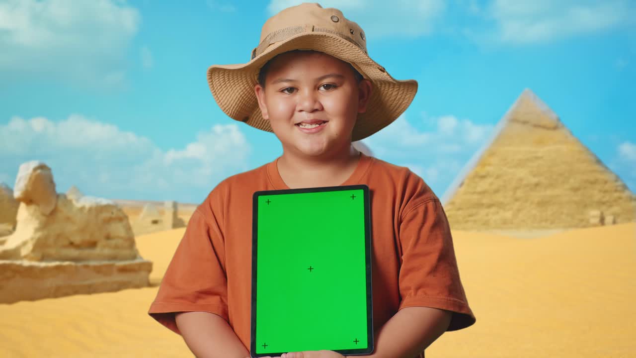 Asian Boy With A Hat And Binoculars Smiling And Showing Green Screen Tablet. Boy Researcher Examines Something While Traveling In Giza Pyramid, Travel Tourism Adventure Concept, Close Up