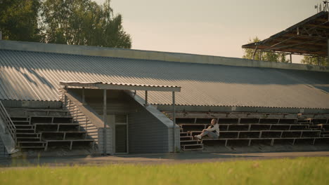 lady seated alone on stadium bleachers in a reflective pose, hands on her knees, background includes metal stadium roof structure with natural trees and greenery