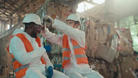 un hombre de piel negra y barba en un uniforme de protección blanco se sienta y se comunica con su colega un hombre moreno con barba y juega a rock-paper-scissors cerca de enormes pilas de basura en una planta de reciclaje y papel residual