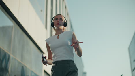 Upper angle view of student dancing with headset on and phone in right hand, moving to music with eyes closed near glass-walled office building