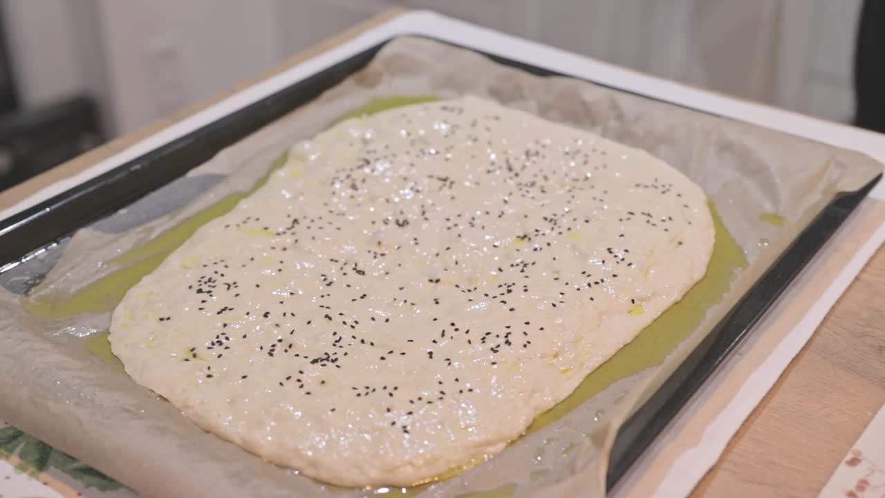 Breve clip di pasta di focaccia cruda che lievita su una teglia, ricoperta di erbe aromatiche e sale marino durante la preparazione in un forno domestico. Movimento naturale di lievitazione. Cottura al forno o processo alimentare in slow motion.